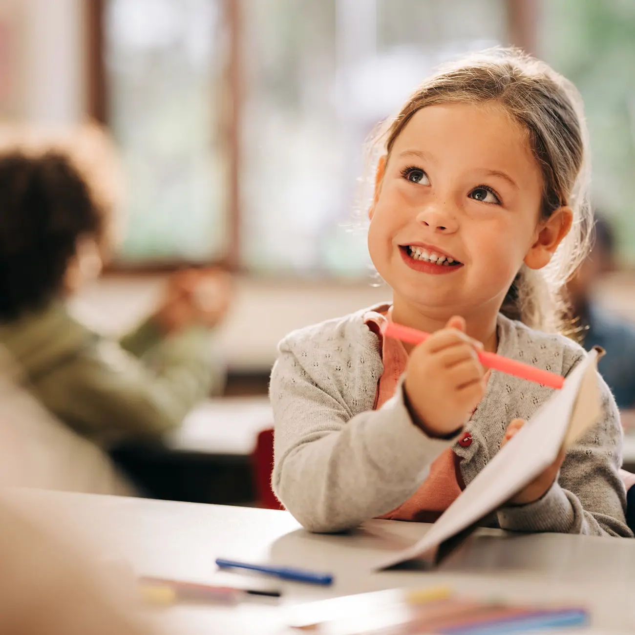 Smiling primary-school girl holding a pink highlighter and notebook, looking up in class