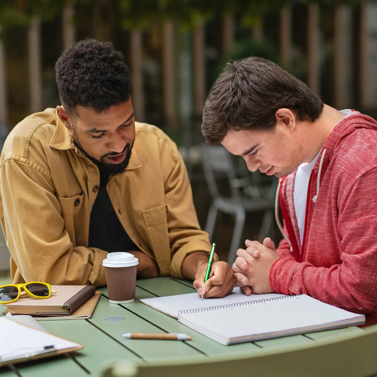 Two students at an outdoor table; one writes in a spiral notebook while the other watches, coffee and notebooks nearby.