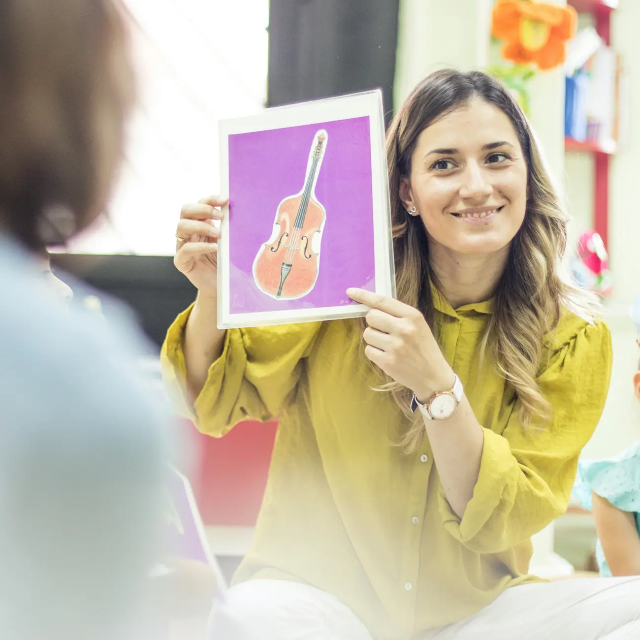 Teacher holding a laminated flashcard showing a cello illustration during a children's music lesson