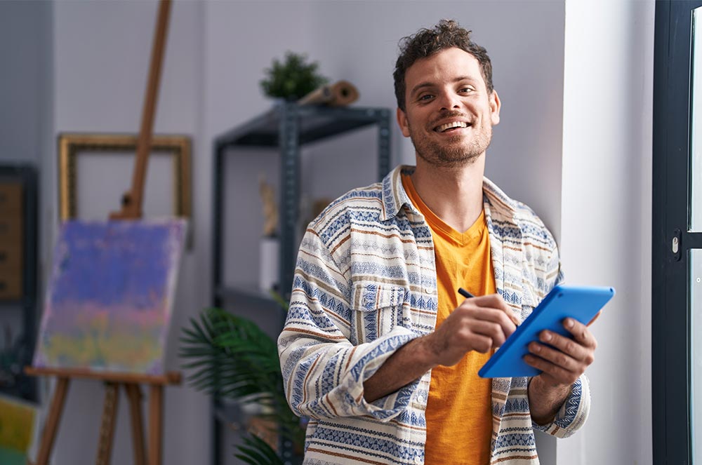 Smiling artist in patterned shirt using a blue tablet in a bright studio with an easel and abstract painting behind