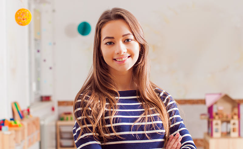 Smiling young woman standing in a nursery classroom with toy shelves and a dolls' house