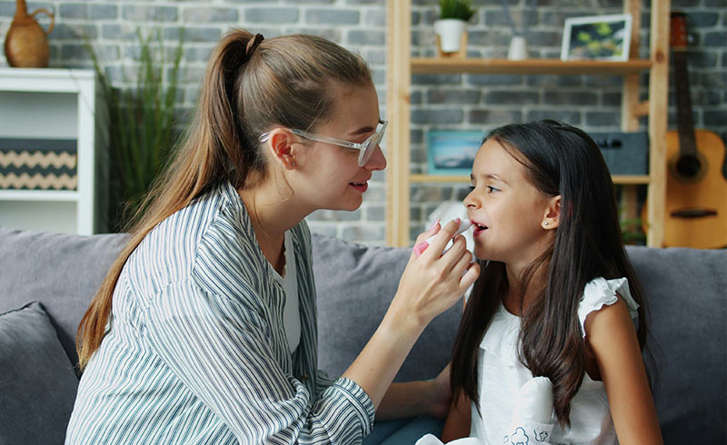 Woman applying lip balm to young girl's lips while they sit together on a living-room sofa