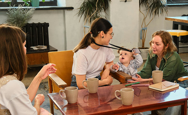 Three women gathered at a table with mugs and notebooks; one holds a baby who reaches for a lanyard