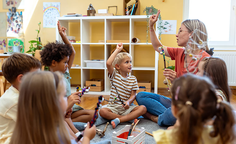 Nursery teacher leading circle time as children sit on the carpet, raise hands and hold decorated craft sticks