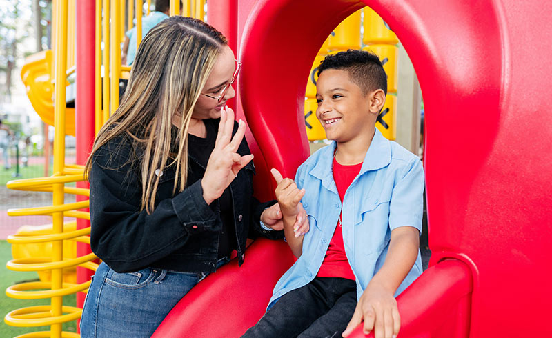 Adult using sign language with smiling boy on red playground slide, practising communication