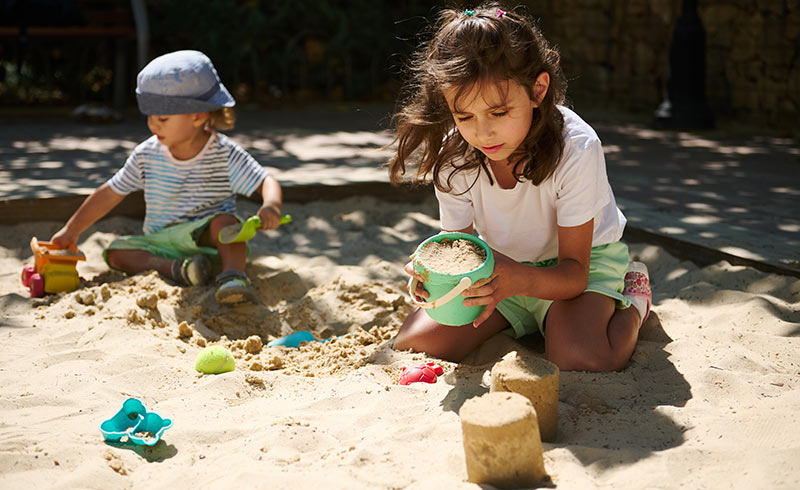 Two children playing in a sandpit - girl filling a green bucket to build sandcastles, younger child with a toy truck