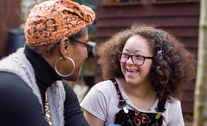 Smiling young woman with glasses and floral dungarees talking with a woman in a patterned headscarf