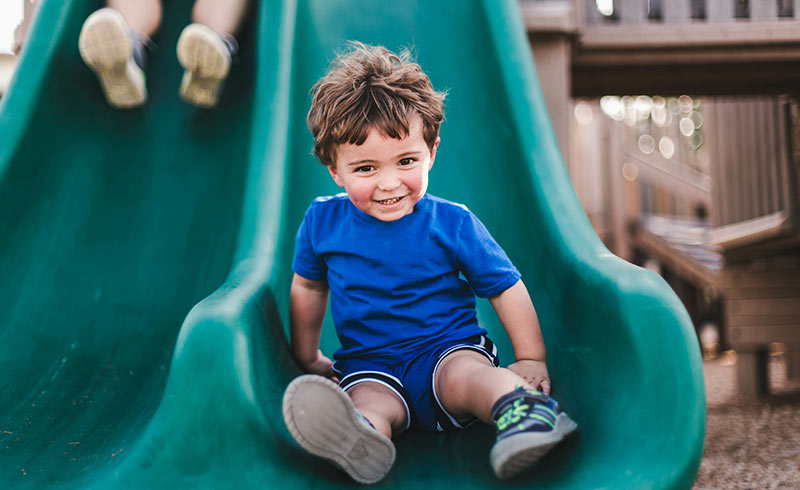 Smiling toddler boy in blue T‑shirt and shorts sliding down a green playground slide