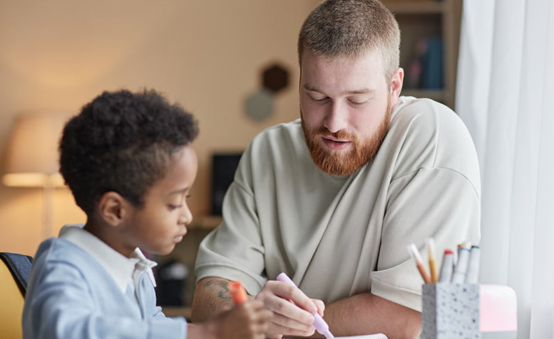 Adult helping a child with homework at a table, using a highlighter with pencils and pens nearby