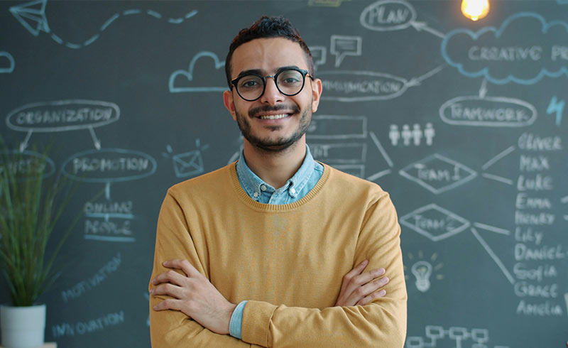 Smiling young man with glasses and folded arms standing before a chalkboard of business diagrams and a team flowchart