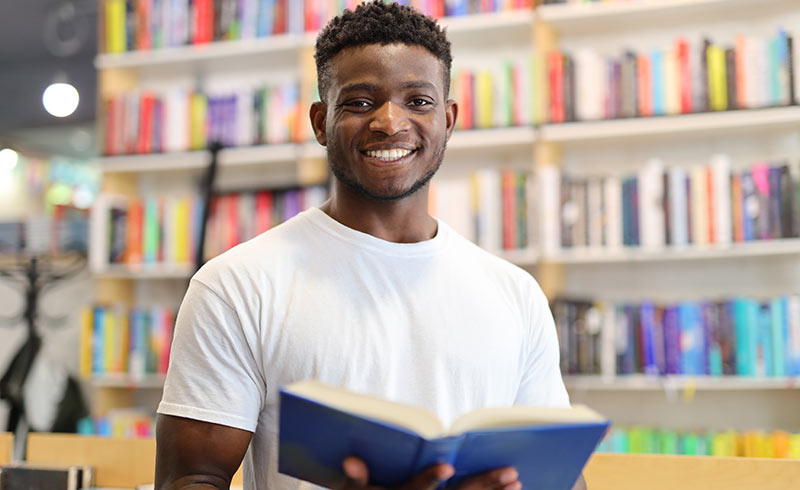 Smiling young man holding an open book in front of colourful bookshelves in a bookshop