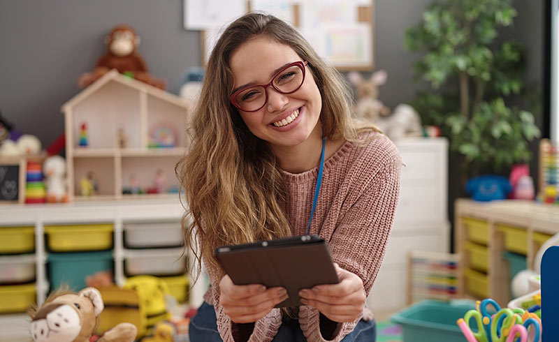 Smiling nursery practitioner wearing a lanyard offering a tablet in a colourful playroom with toys and storage