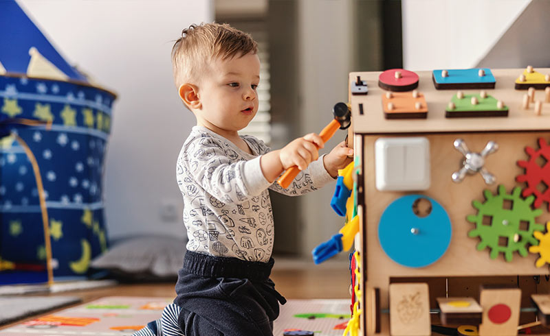 Toddler playing with a wooden activity cube, using a small hammer to explore gears and shape pegs