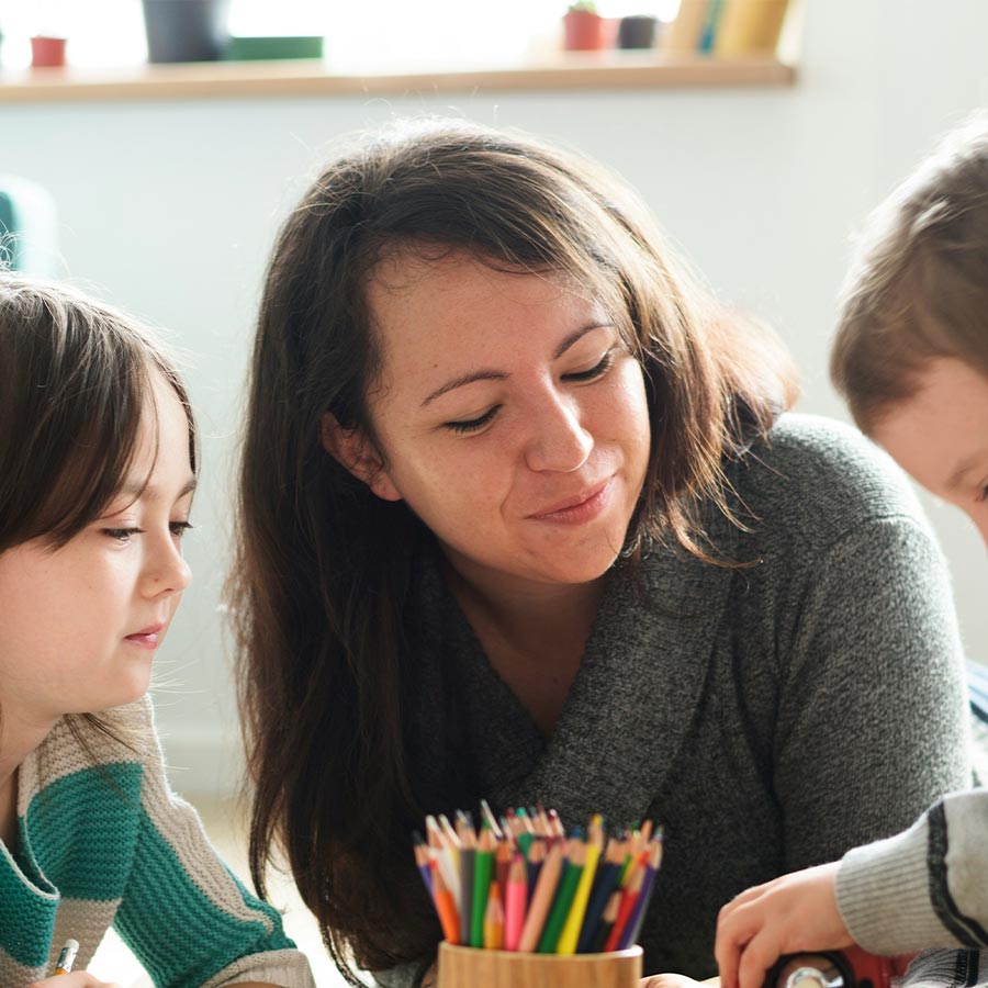 Woman helping two children draw at a table; pot of coloured pencils visible