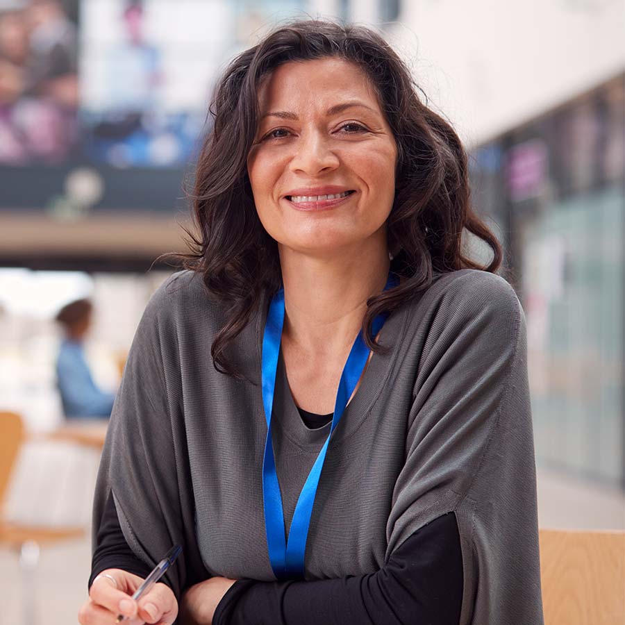Smiling woman with blue lanyard holding a pen, seated in a bright conference or campus lobby