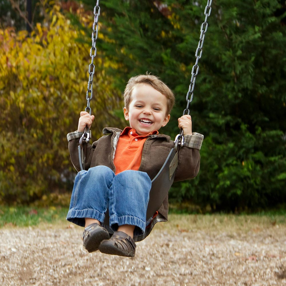 Smiling young child in orange top and brown jacket swinging on chained seat in a park with autumn foliage