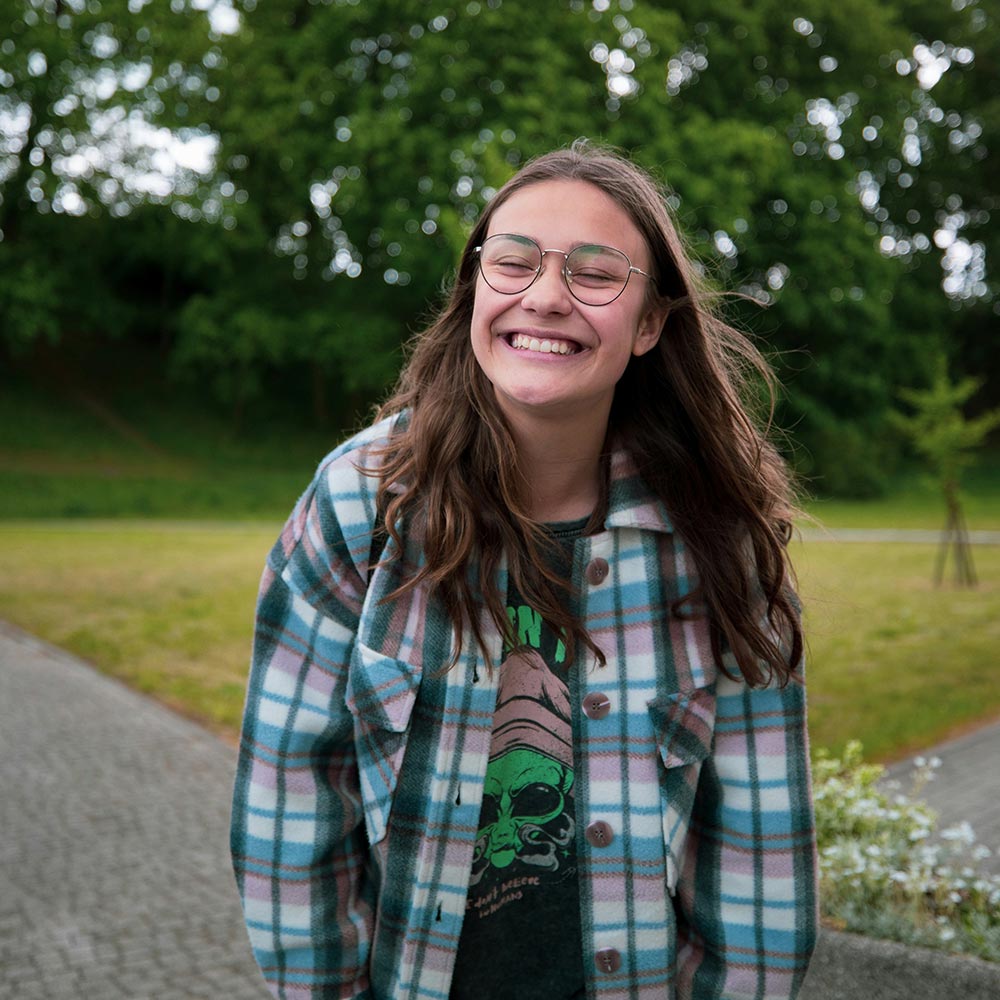 Young person smiling broadly, wearing round glasses, a green‑blue checked jacket and a T‑shirt with a green skull graphic in a park