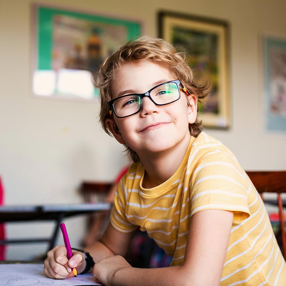 Smiling pupil with glasses in yellow striped T-shirt, drawing with a pink pencil at a classroom desk