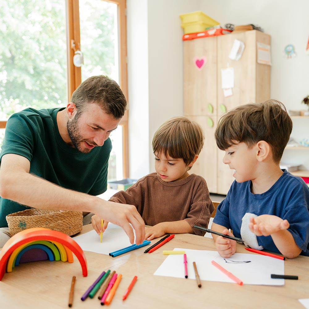 Adult guiding two young boys drawing with coloured pencils at a table, wooden rainbow toy and papers visible