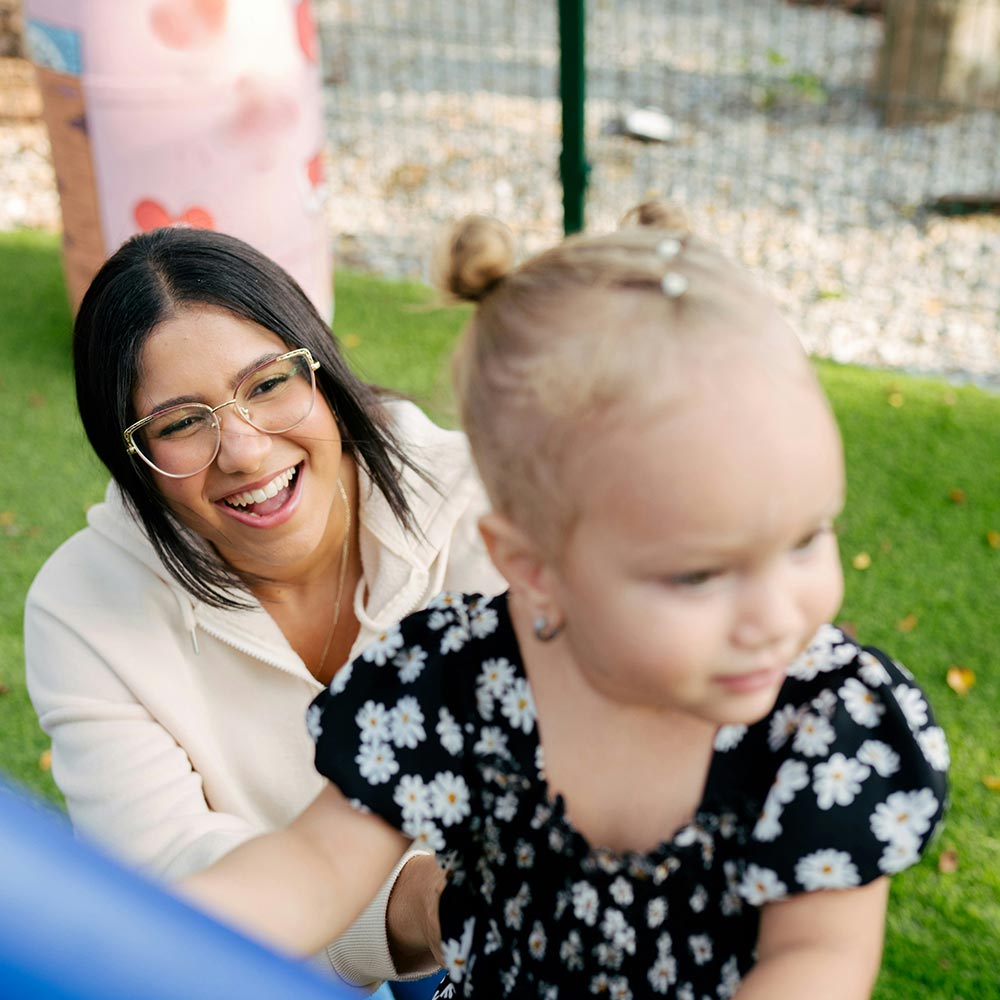 Smiling carer helping a toddler in a black daisy-print dress climb playground equipment on grass