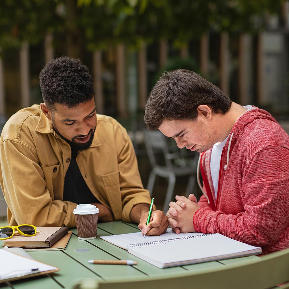 Tutor pointing with pencil at a spiral notebook while a student writes at an outdoor table, takeaway coffee beside them