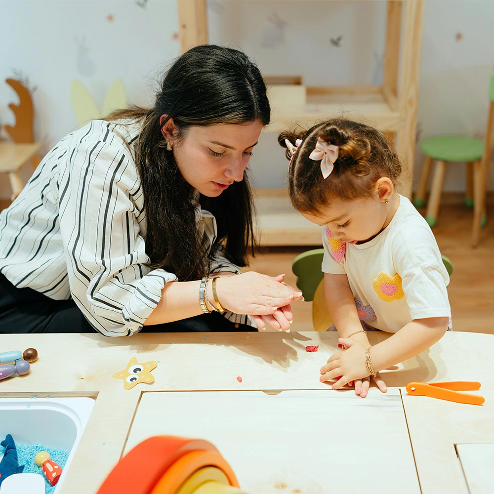 Carer guiding a toddler's hands as they shape playdough at a nursery activity table