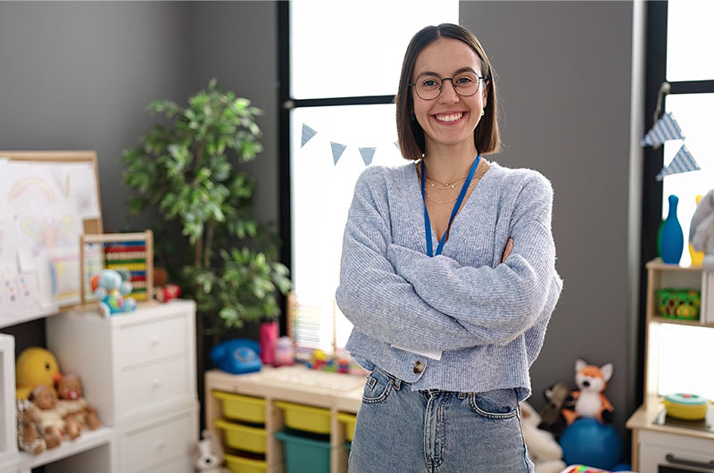 Preschool teacher smiling, arms crossed, wearing a lanyard in a colourful classroom with toys.